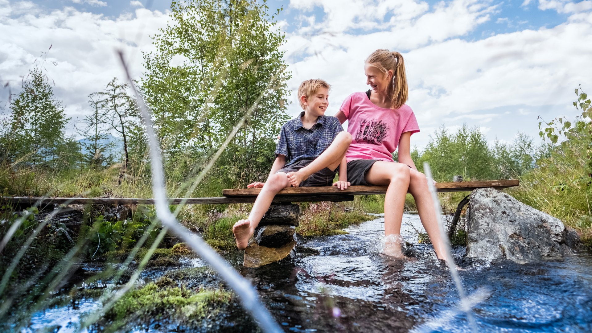 Smaragdhotel Tauernblick – Ihr 4-Sterne-Hotel am Wildkogel Zwei Kinder sitzen auf einer Holzbank über einem Bach im Freien